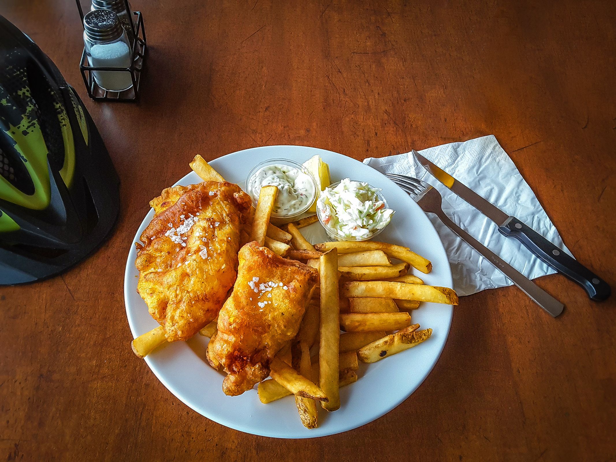 A plate of fish and chips with coleslaw and tartar sauce sits on a wooden table next to a bicycle helmet.