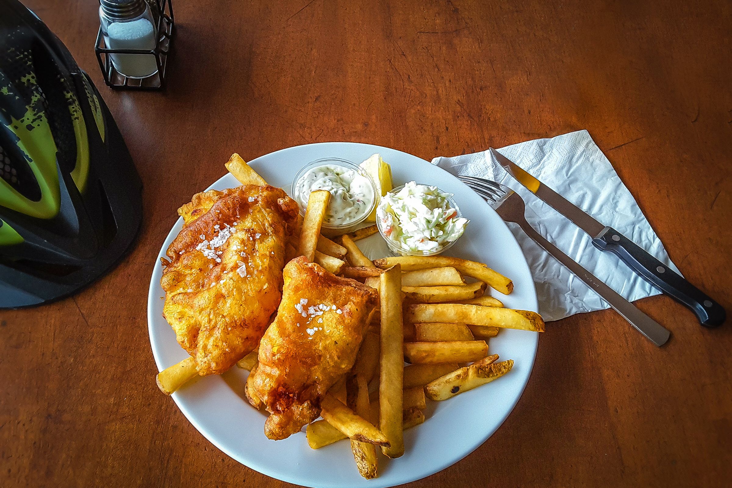 A plate of fish and chips with coleslaw and tartar sauce sits on a wooden table next to a bicycle helmet.