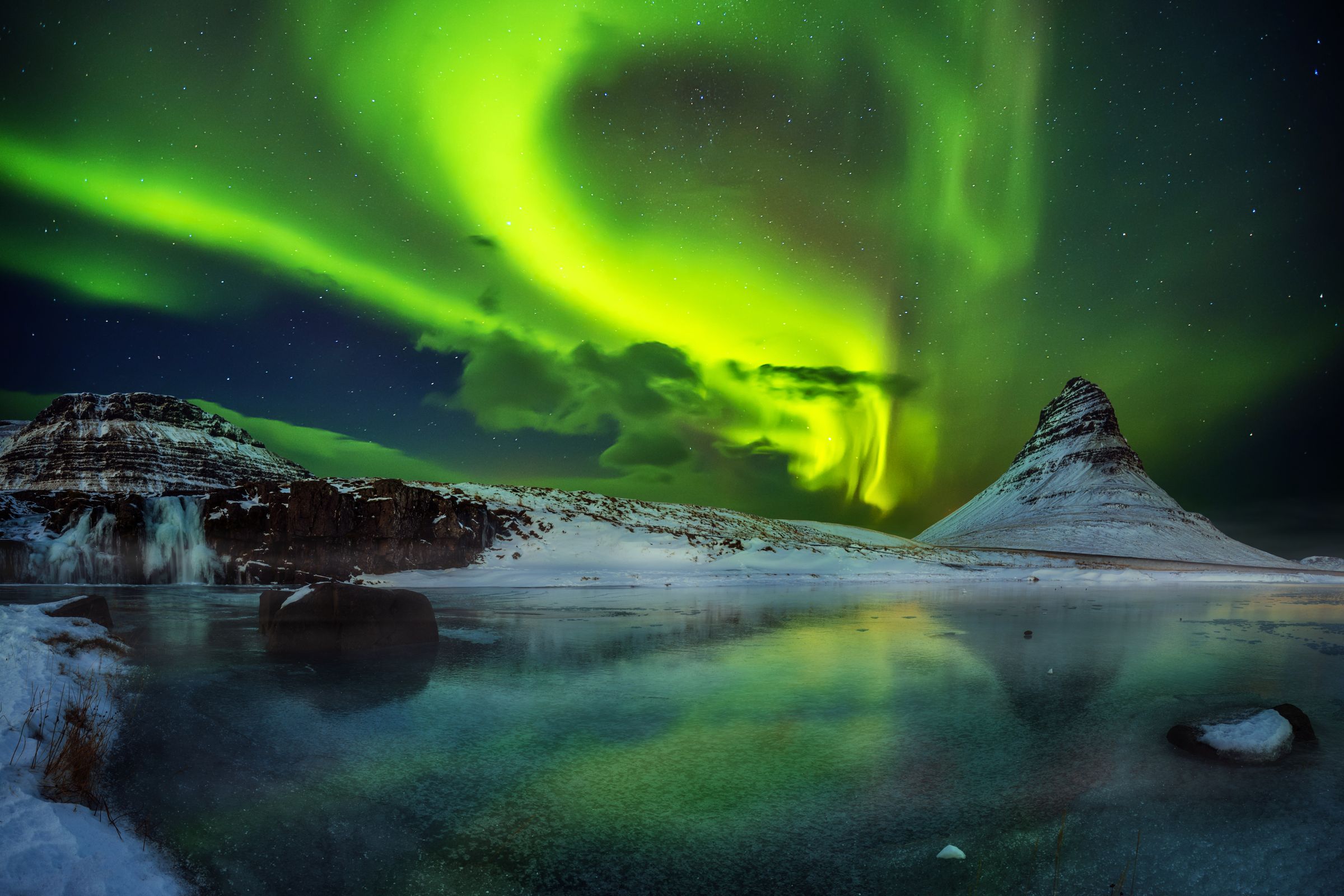Unas vibrantes auroras boreales verdes se arquean sobre una montaña cónica cubierta de nieve y un lago en calma que refleja sus luces.