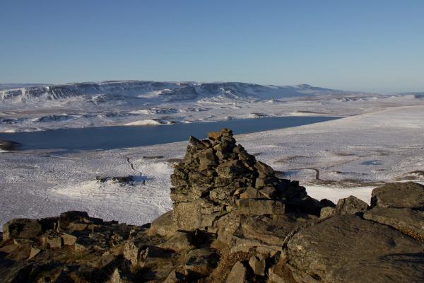 A rocky cairn overlooks a snow-covered landscape with a lake and distant mountains under a clear blue sky.