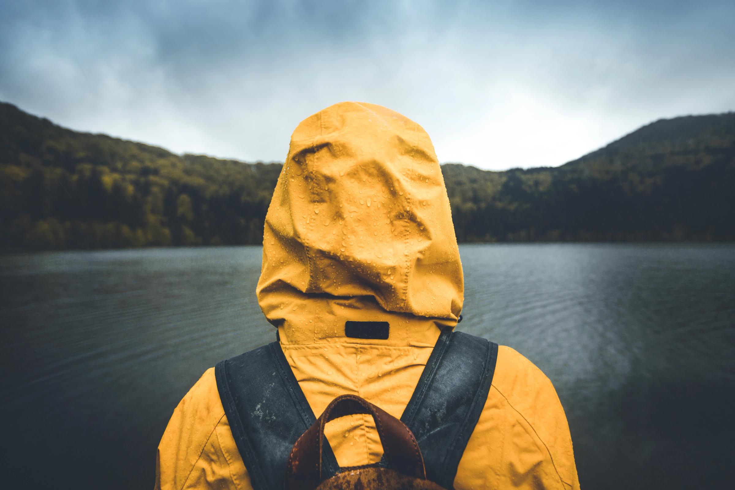 A person in a yellow rain jacket and backpack looks out at a lake and mountains on a rainy, overcast day.