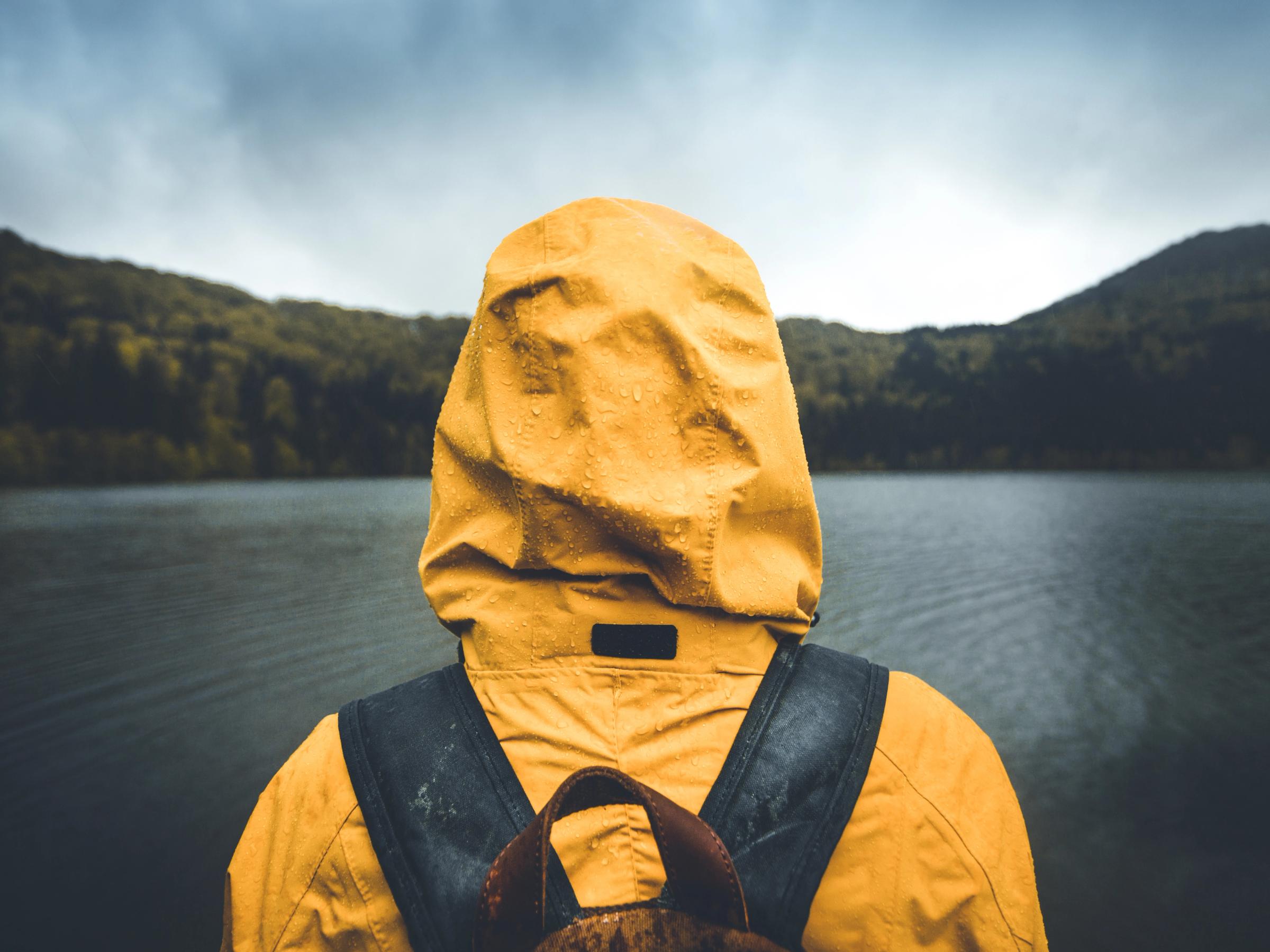 Person in a yellow hooded raincoat with water droplets and backpack looks out at a lake and forested hills under a cloudy sky.