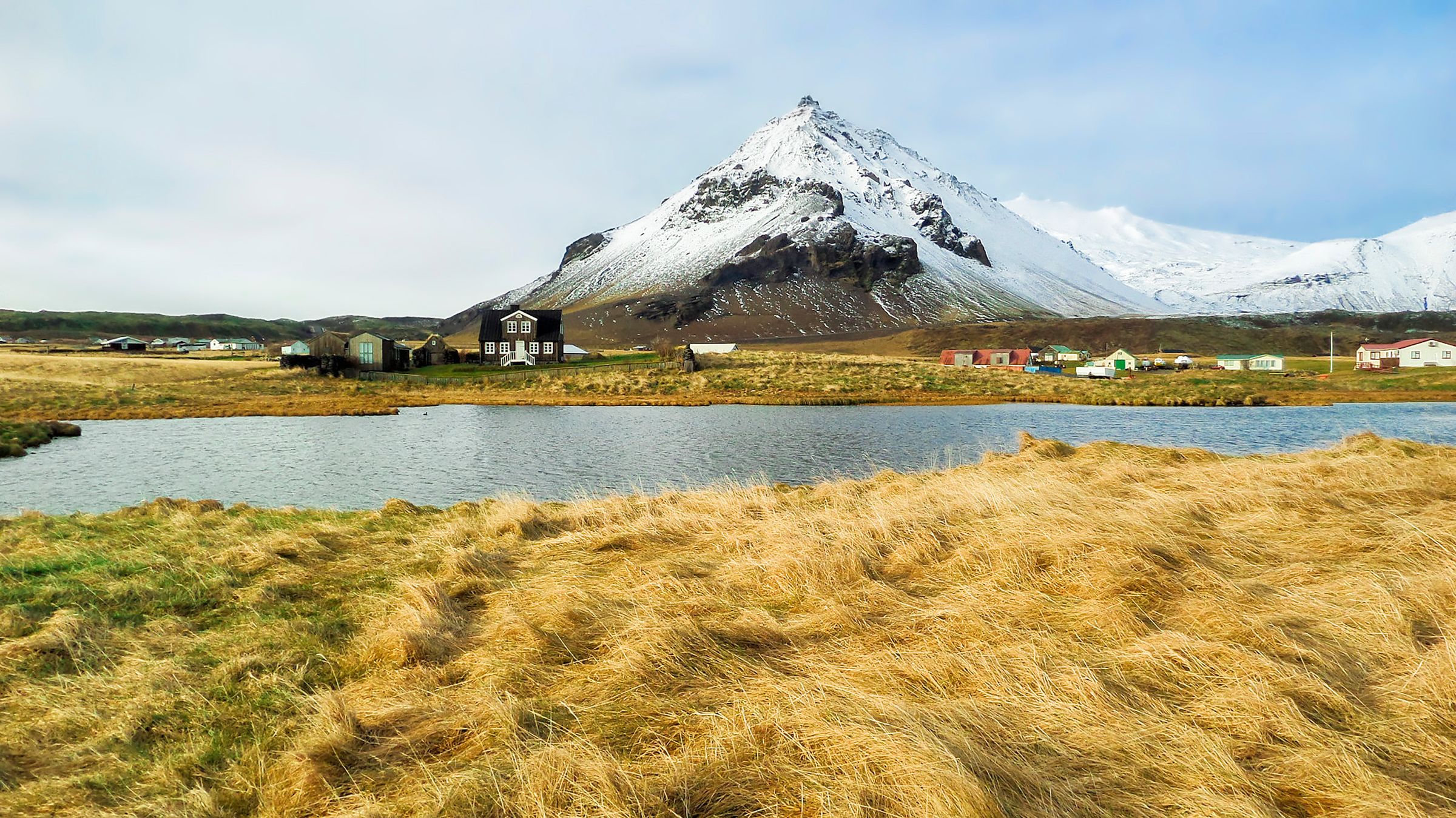Campos dorados y un lago en primer plano, con una casa oscura, un pequeño pueblo colorido y una gran montaña nevada en el fondo.