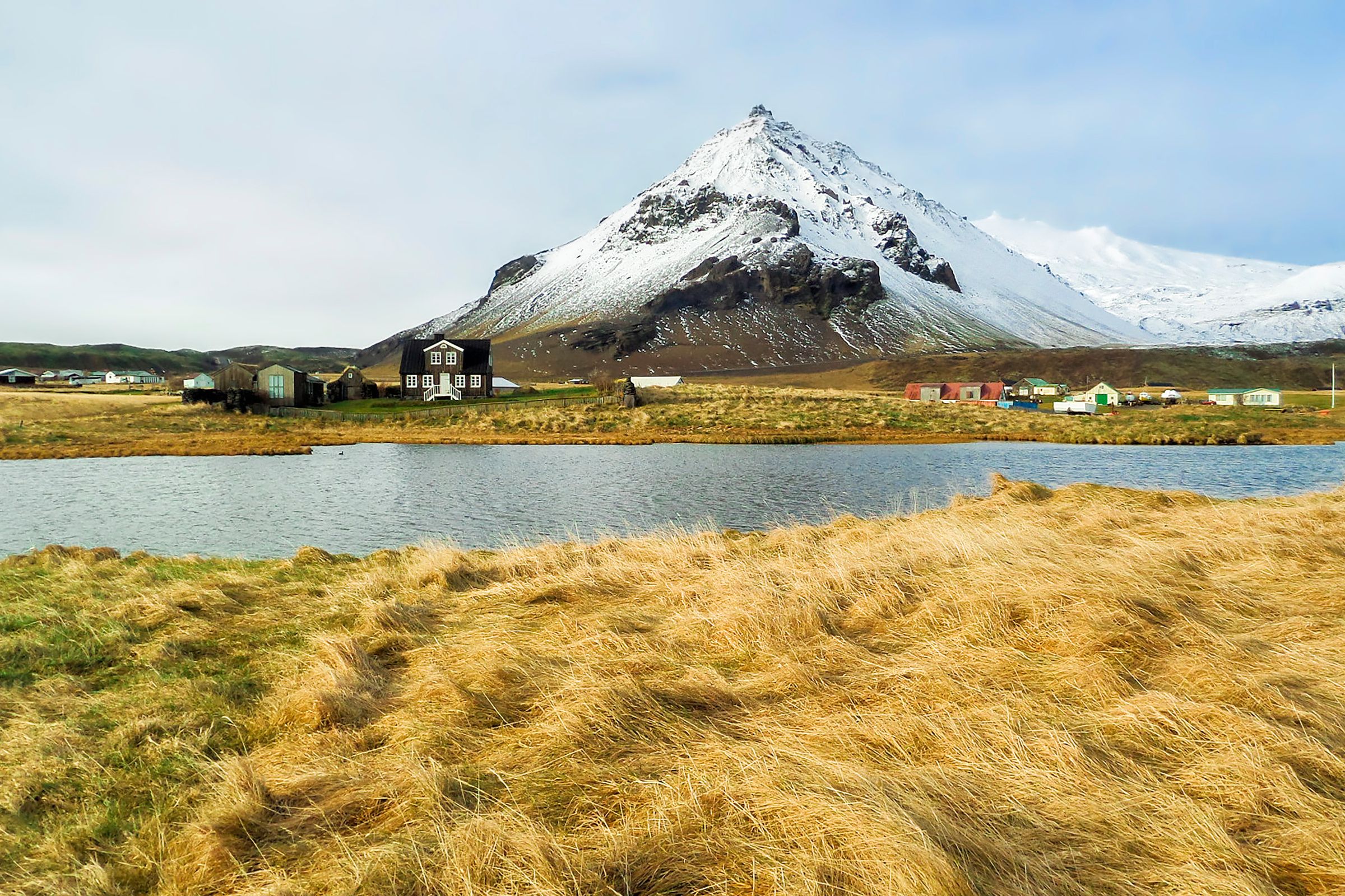 Campos dorados y un lago en primer plano, con una casa oscura, un pequeño pueblo colorido y una gran montaña nevada en el fondo.