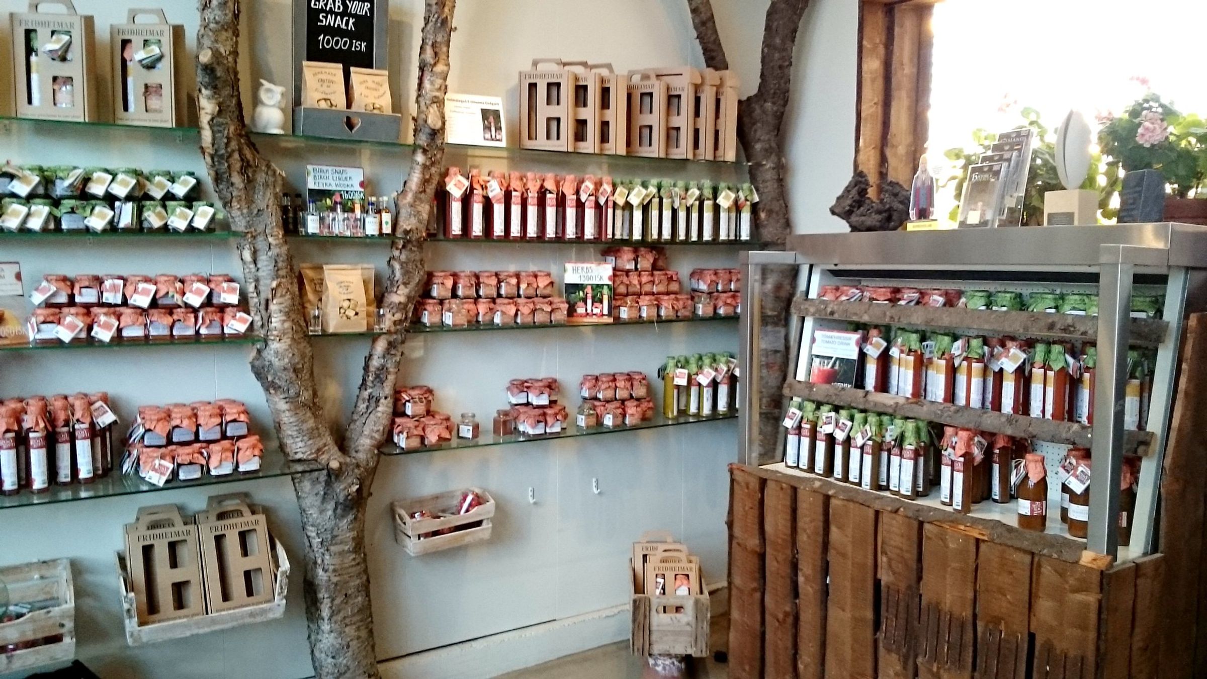 Interior de una tienda rústica con estanterías de cristal construidas alrededor de un tronco de árbol, donde se exhiben numerosos frascos y botellas con productos alimenticios.