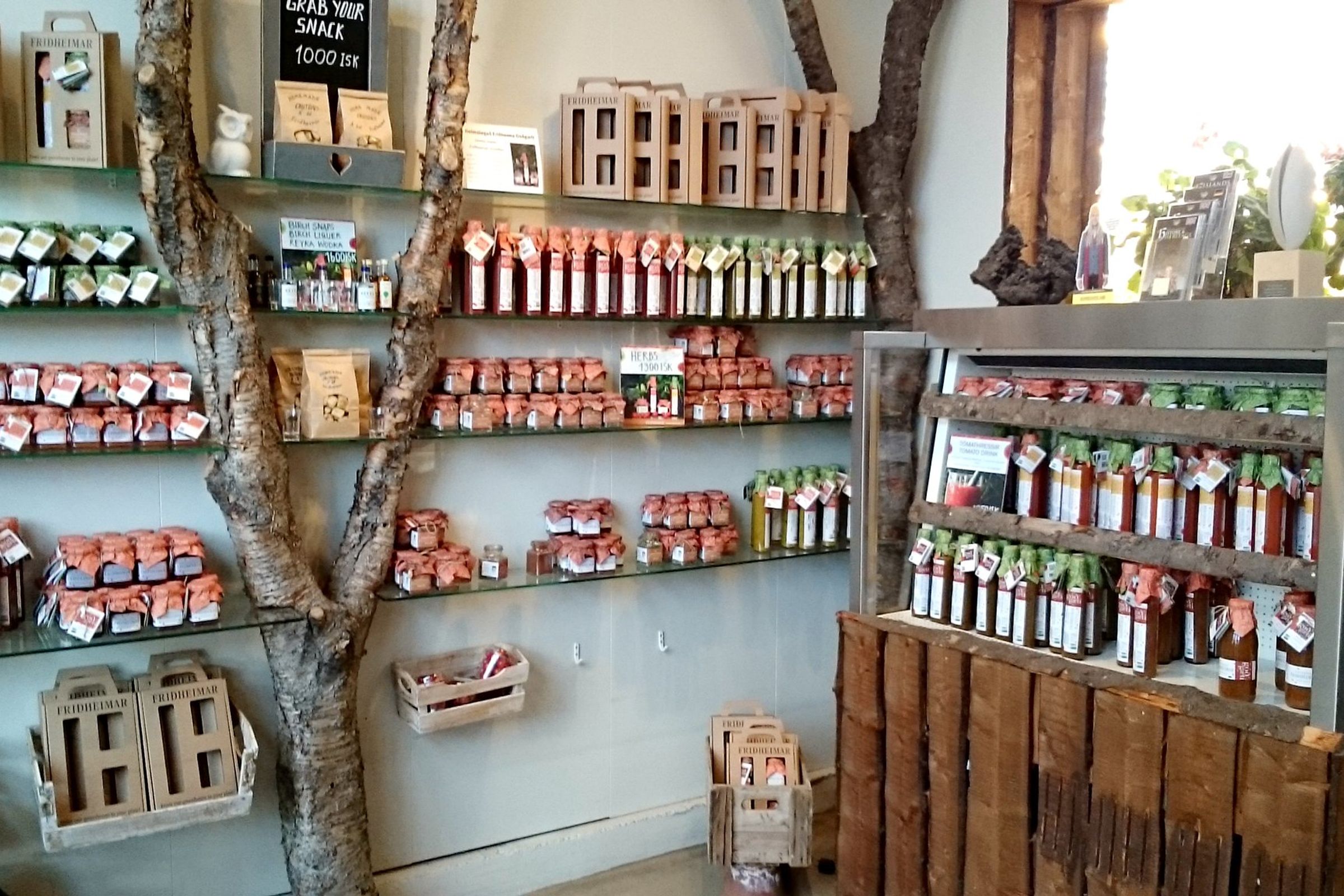 Interior de una tienda rústica con estanterías de cristal construidas alrededor de un tronco de árbol, donde se exhiben numerosos frascos y botellas con productos alimenticios.