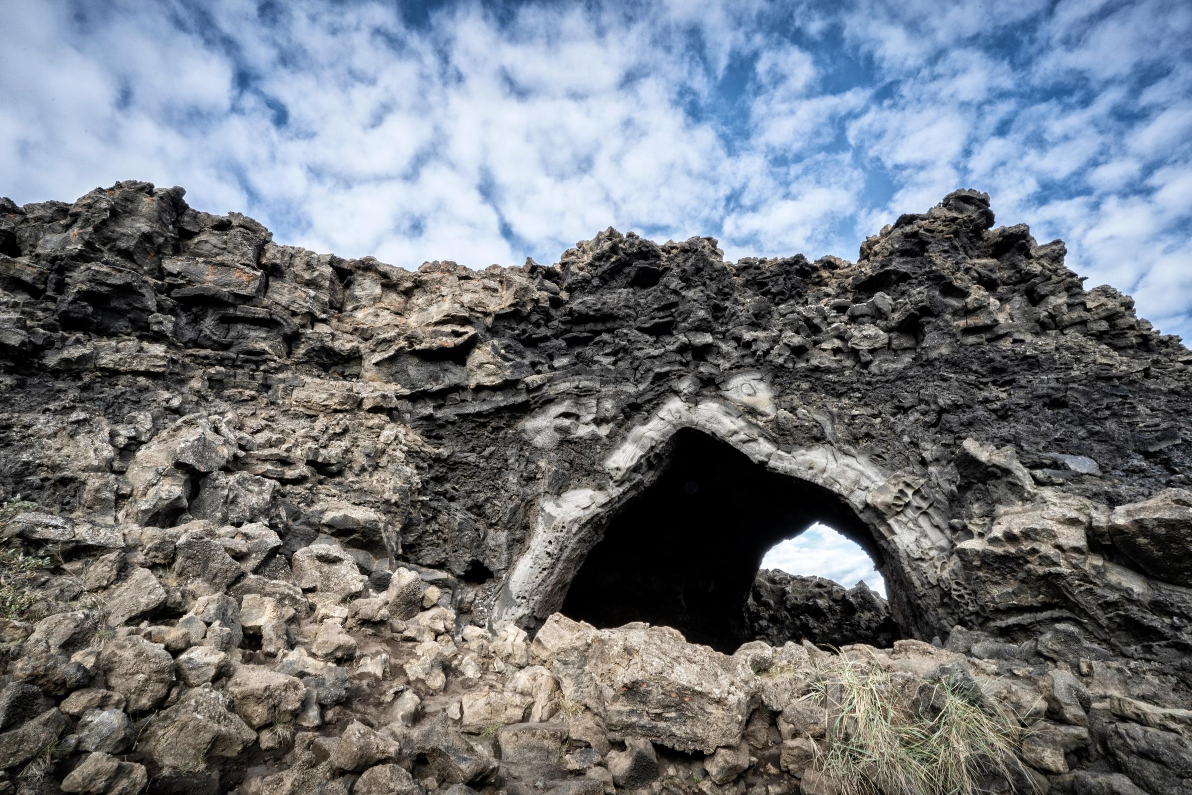Arco natural en oscura y escarpada roca volcánica bajo un cielo azul nublado.