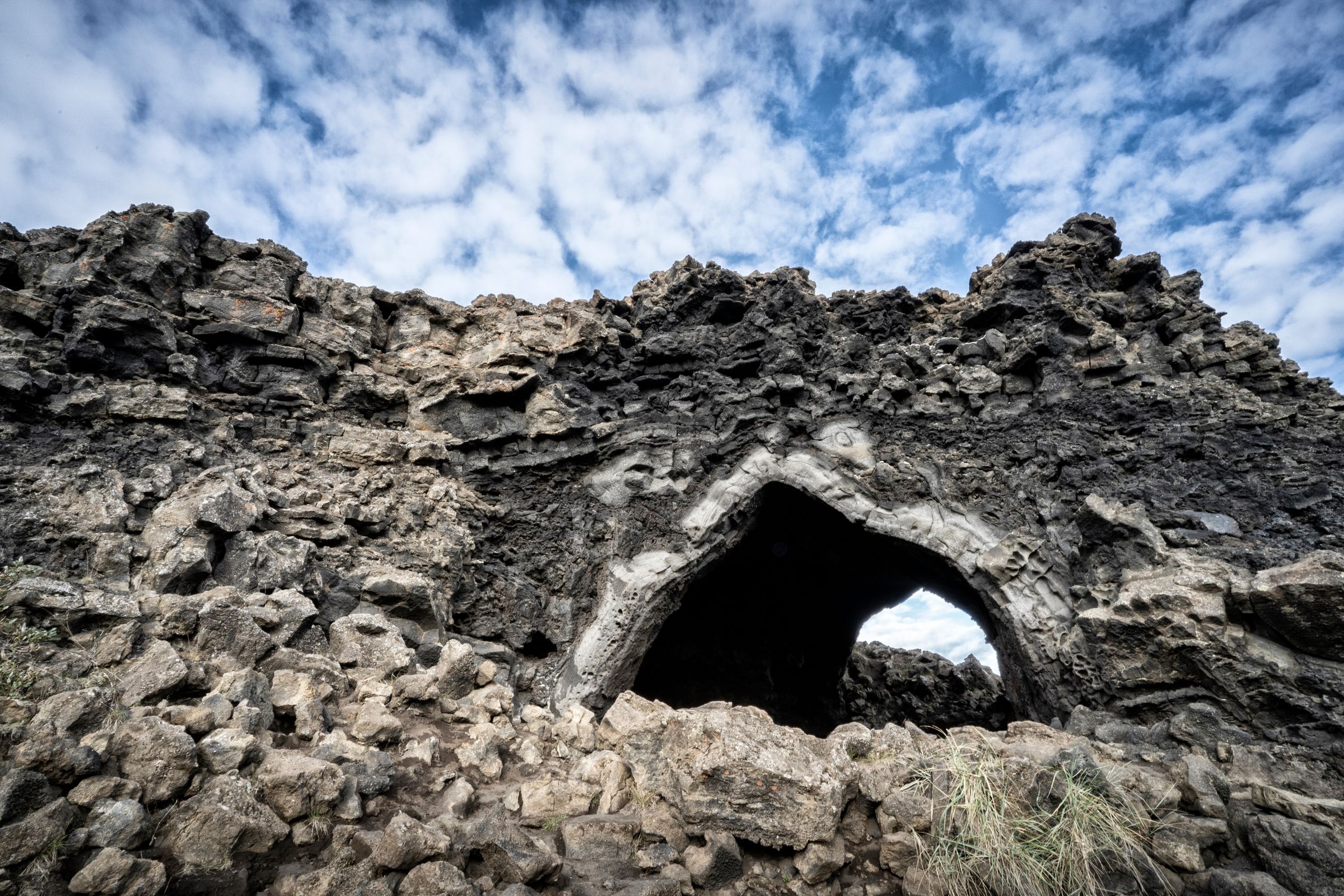Arco natural en oscura y escarpada roca volcánica bajo un cielo azul nublado.