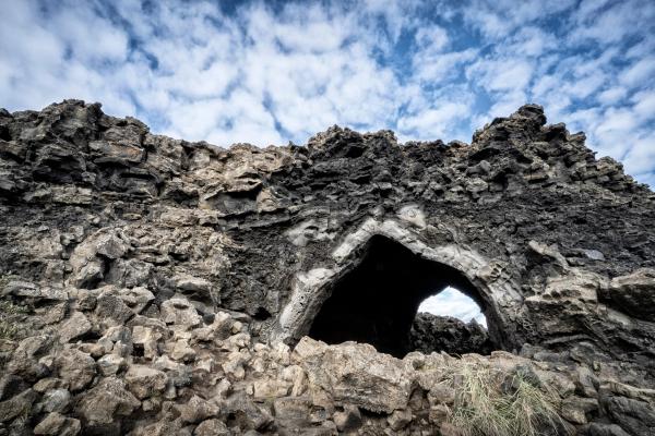Natural archway in dark, rugged volcanic rock under a cloudy blue sky.