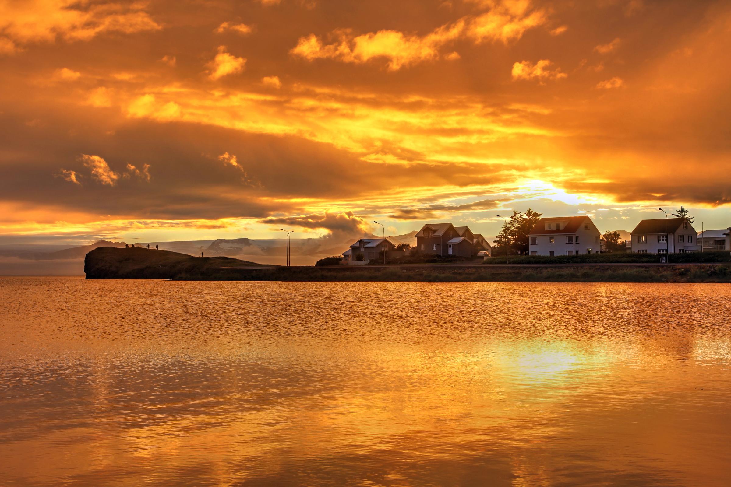 Puesta de sol vibrante de color naranja reflejándose sobre el agua tranquila, con casas y un afloramiento rocoso en la orilla distante.