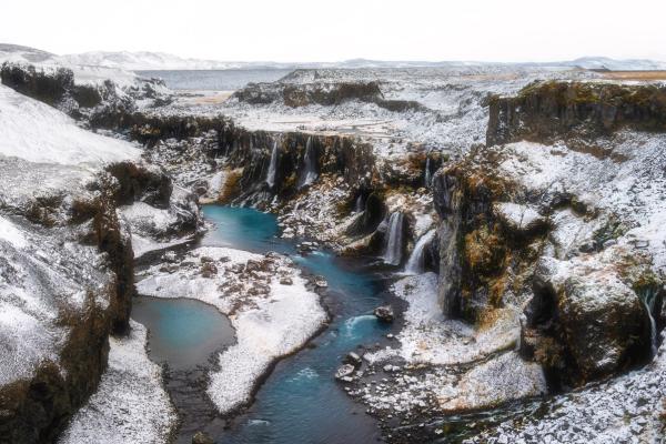 A vibrant blue river flows through a snow-covered canyon with waterfalls.