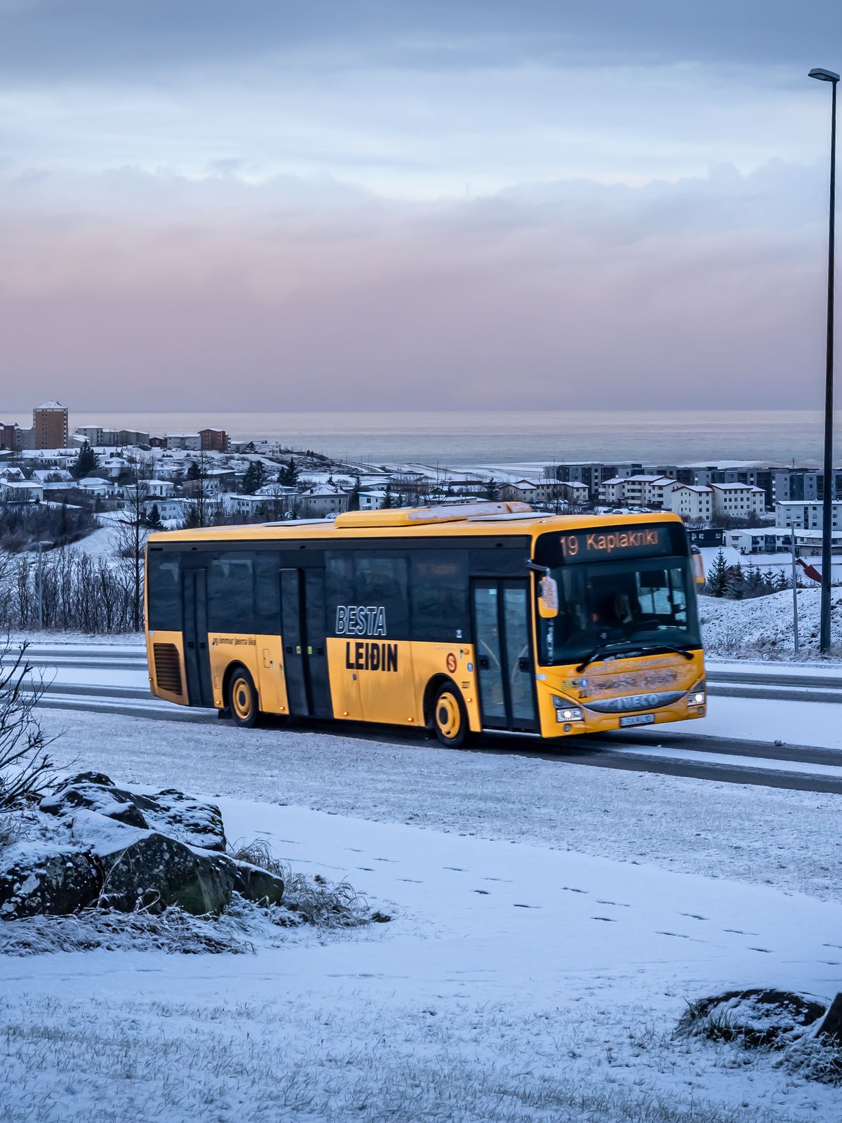 A yellow bus drives on a snowy road with a coastal town in the distance.