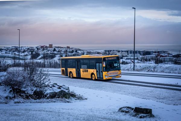 A yellow bus drives on a snowy road, past a town and sea under a cloudy sky.