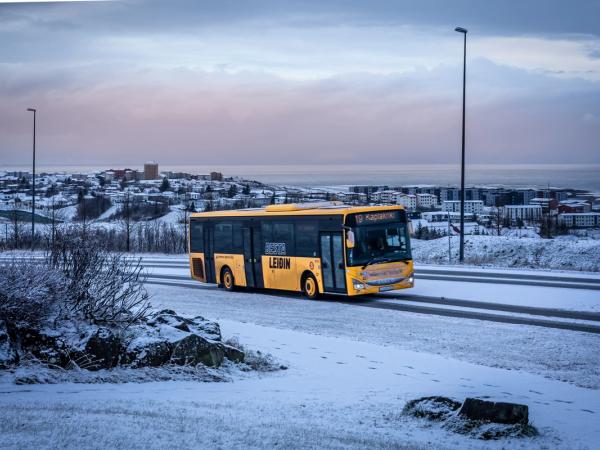 Un autobús amarillo circula por una carretera nevada con un pueblo y el mar de fondo bajo un cielo nublado.