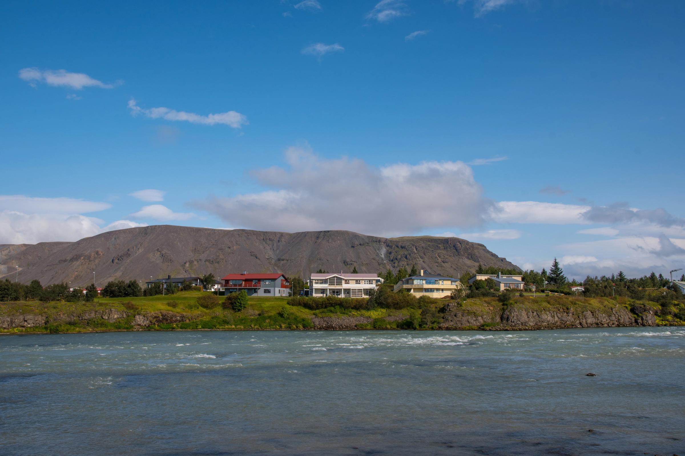 A river flows past a green bank with houses, backed by a dark mountain under a blue sky.