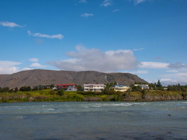 A river flows past a green bank with houses, backed by a dark mountain under a blue sky.