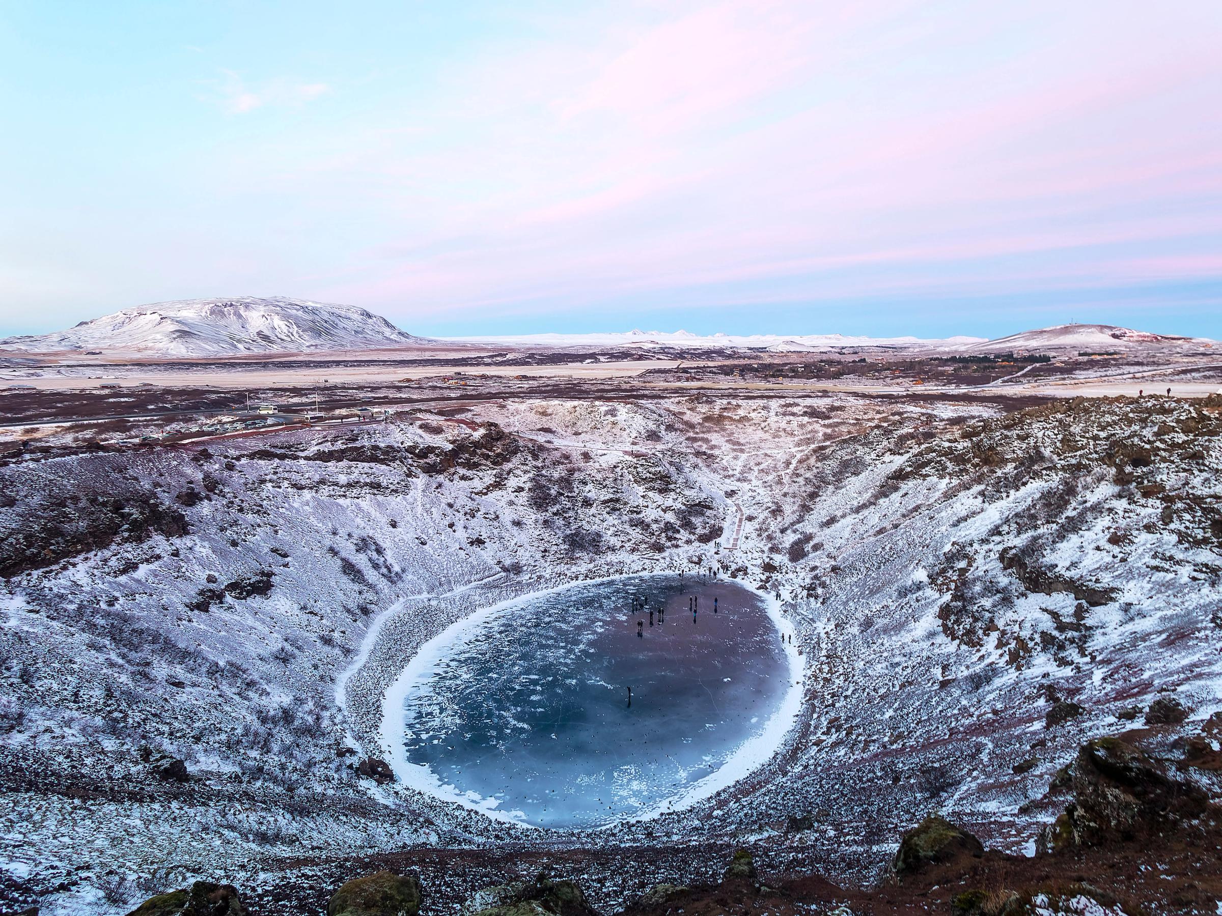 Un cráter volcánico nevado con un lago azul congelado y gente sobre el hielo bajo un cielo rosa y azul.