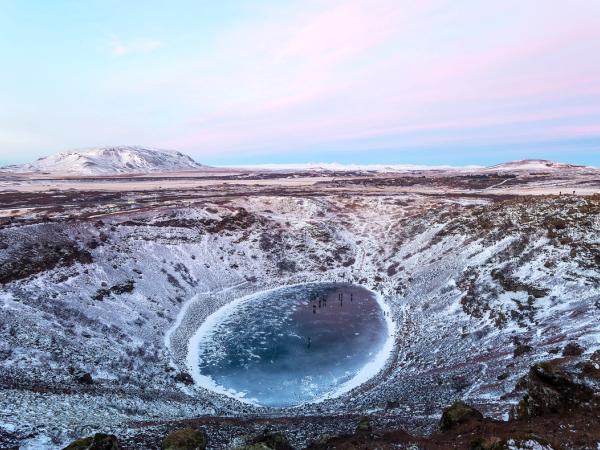 A snowy volcanic crater with a frozen blue lake and people on the ice under a pink and blue sky.