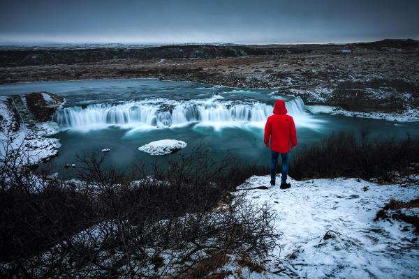 A person in a red jacket stands on a snowy bank overlooking a long-exposure waterfall.