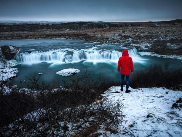 Person in red jacket views misty waterfall in a snowy landscape.