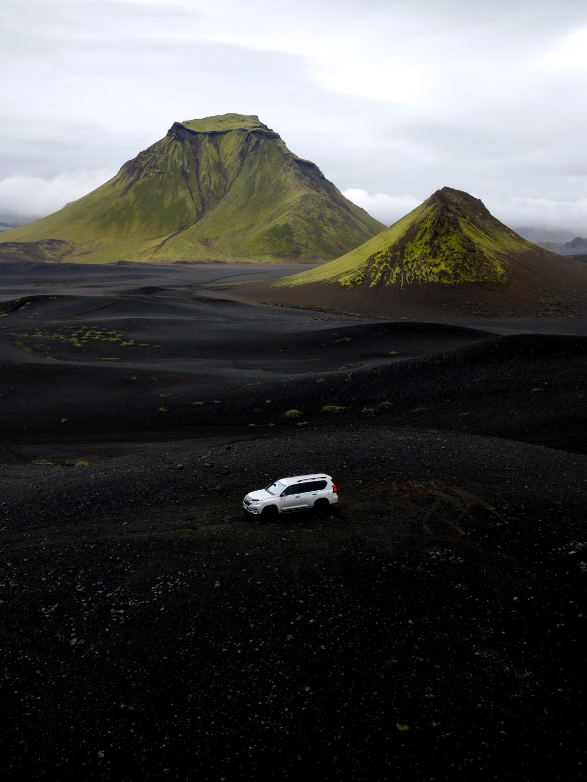 White SUV on a black volcanic landscape with green mountains under a cloudy sky.