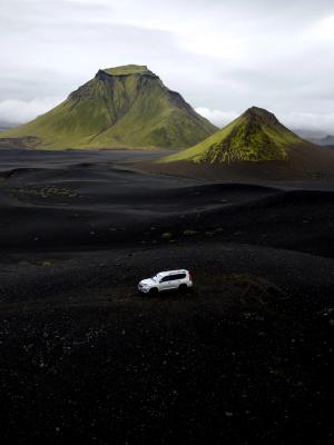 White SUV on a black volcanic landscape with green mountains under a cloudy sky.