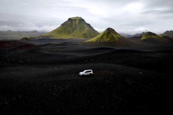 White SUV on black volcanic plains with green mountains under a cloudy sky.