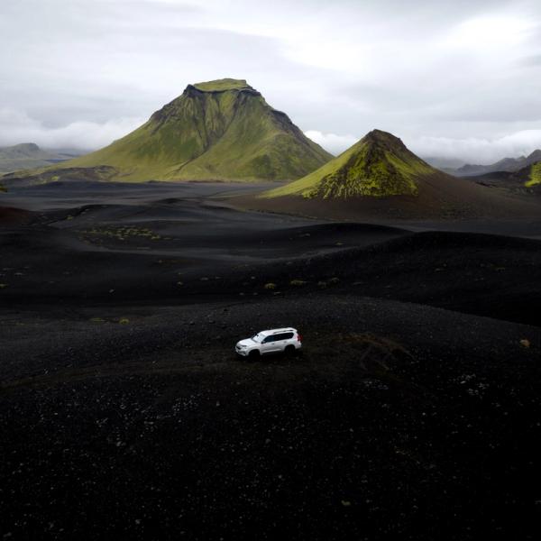 White SUV on a black landscape with green, moss-covered mountains under a cloudy sky.
