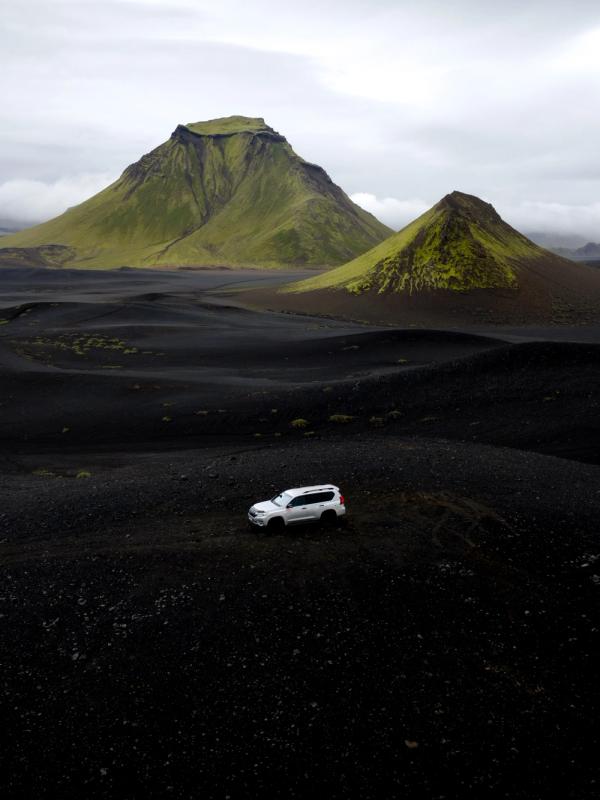 White SUV on a black volcanic landscape with green mountains under a cloudy sky.