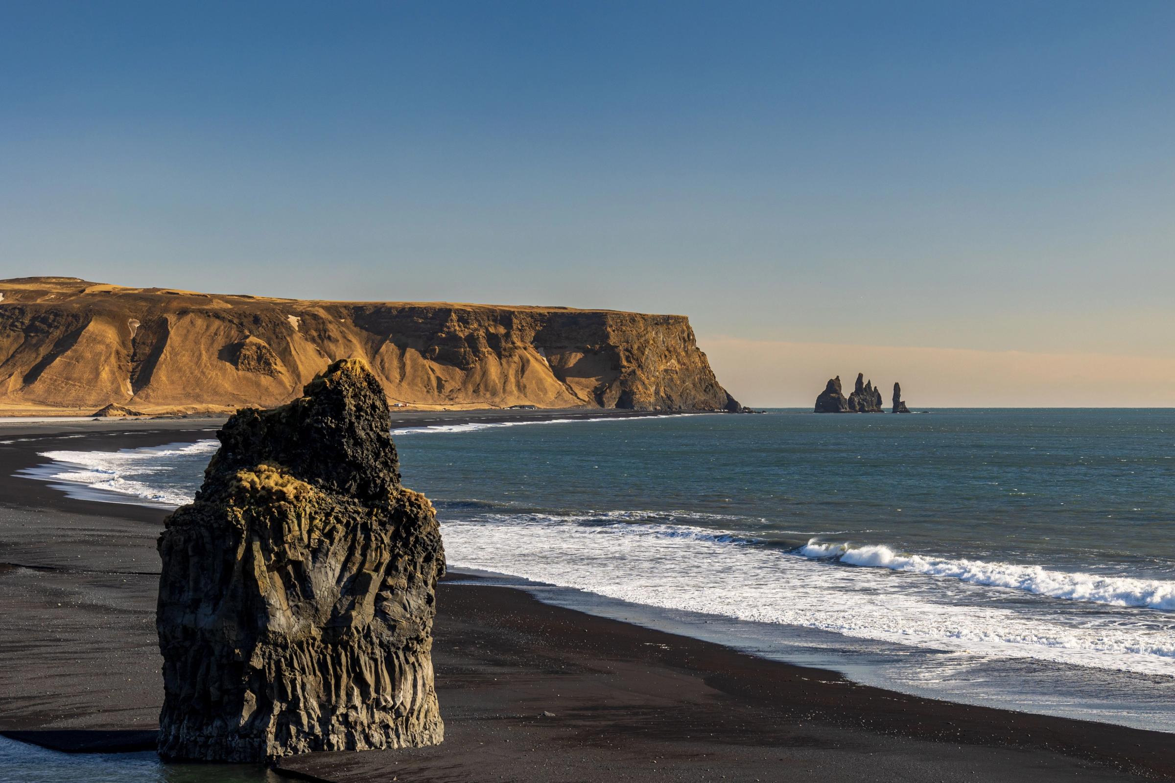 Playa de arena negra con una gran roca oscura, olas del océano, acantilados y columnas de basalto en el mar bajo un cielo azul.