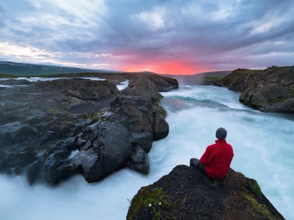 a man is sitting on a rock overlooking a waterfall at sunset in iceland.