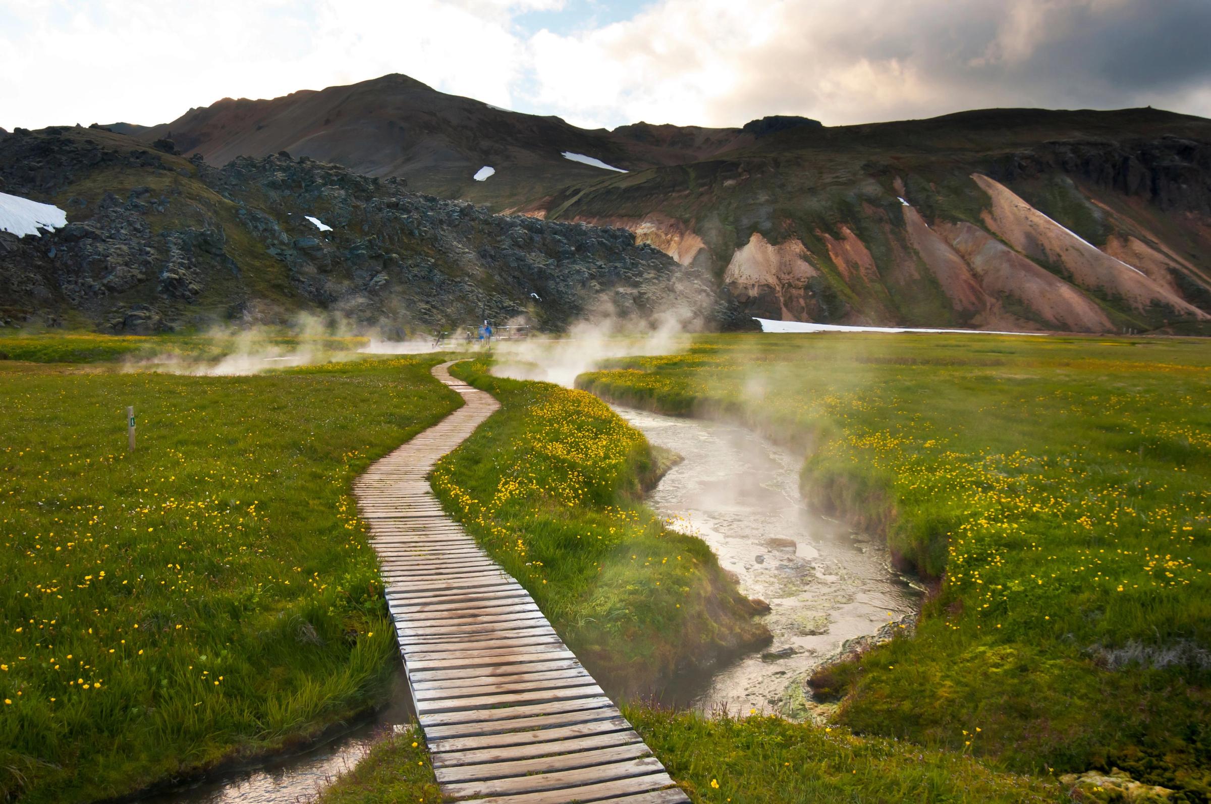 Aerial view of a hot spring in Iceland with a wooden boardwalk and snow covered mountains in the background