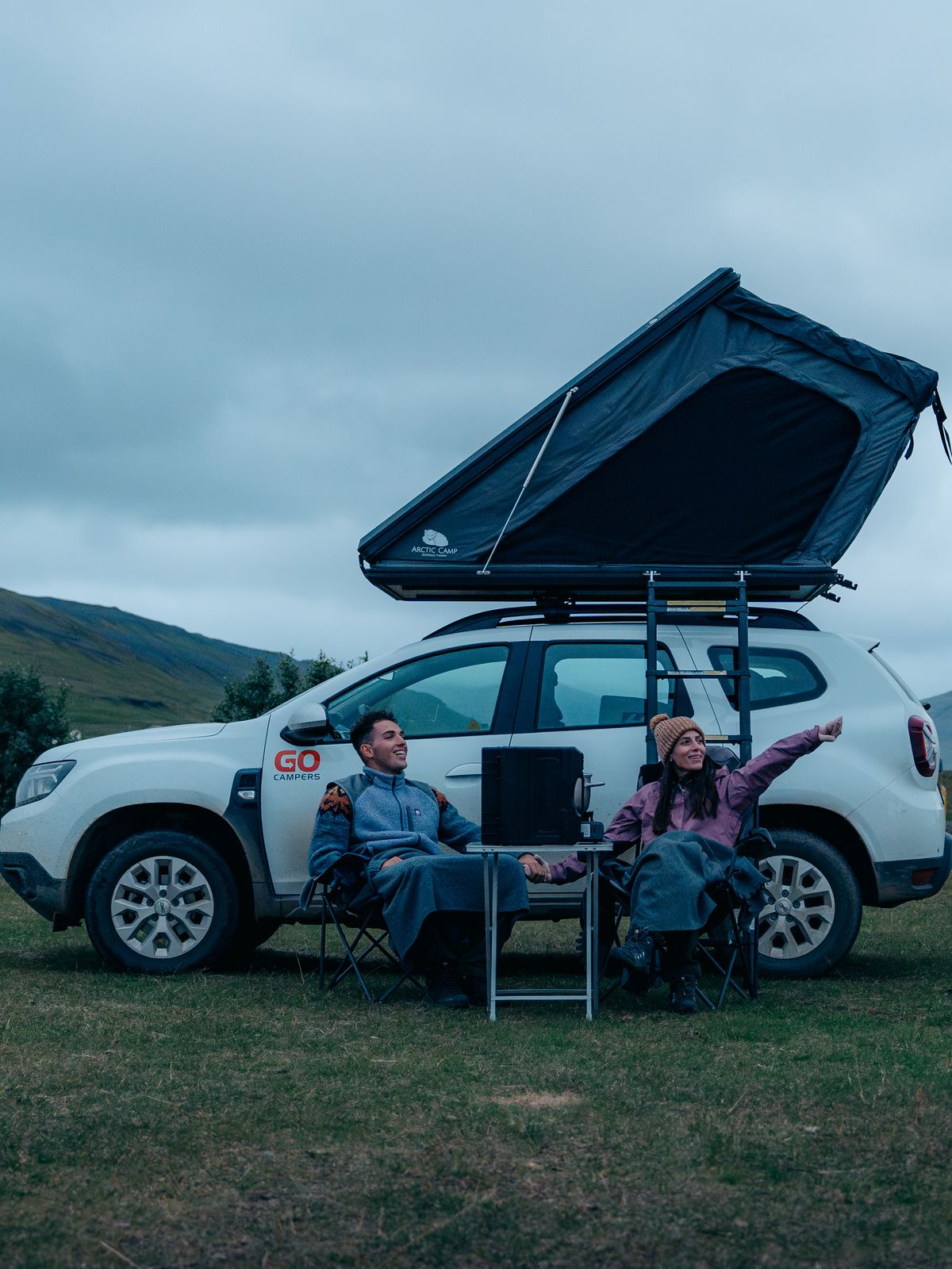 a man and a woman are sitting in front of a car with a tent on top of it .