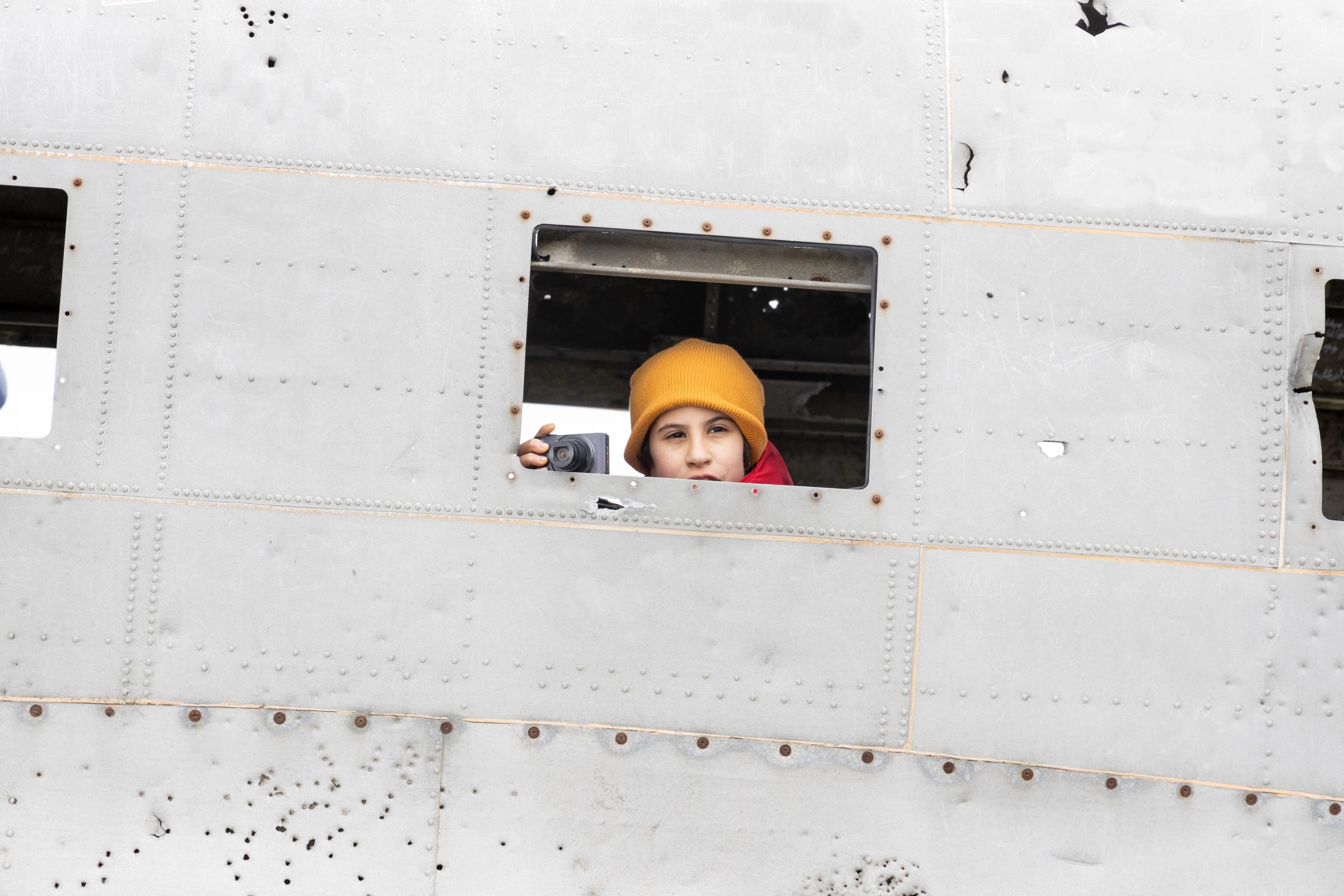 Child in a yellow hat looks out a window of a riveted metal structure, holding a camera.