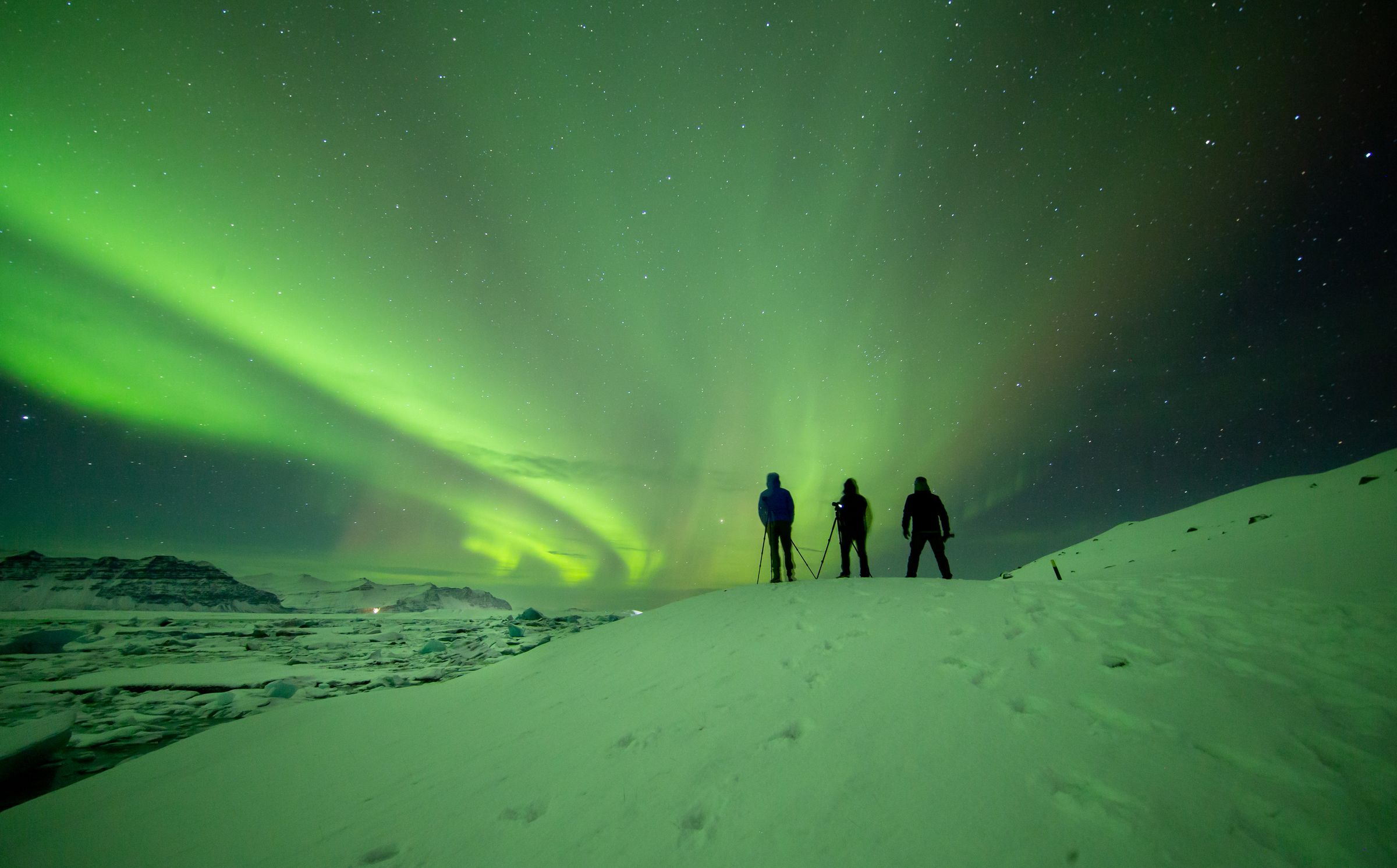 three people are standing on top of a snow covered hill watching the aurora borealis .