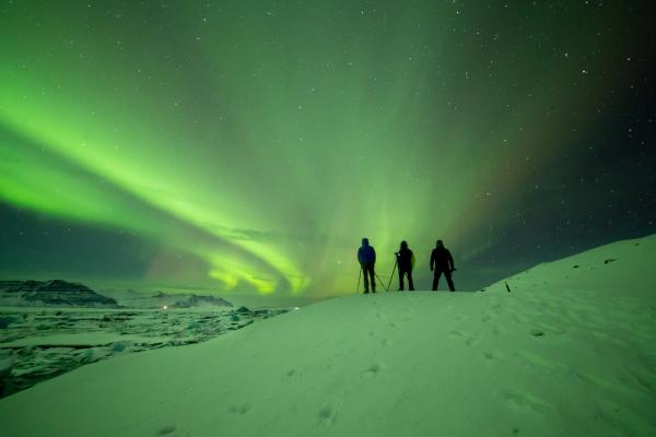 three people are standing on top of a snow covered hill watching the aurora borealis .