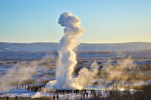 A tall column of steam erupts from a geyser, surrounded by spectators in a snowy landscape.