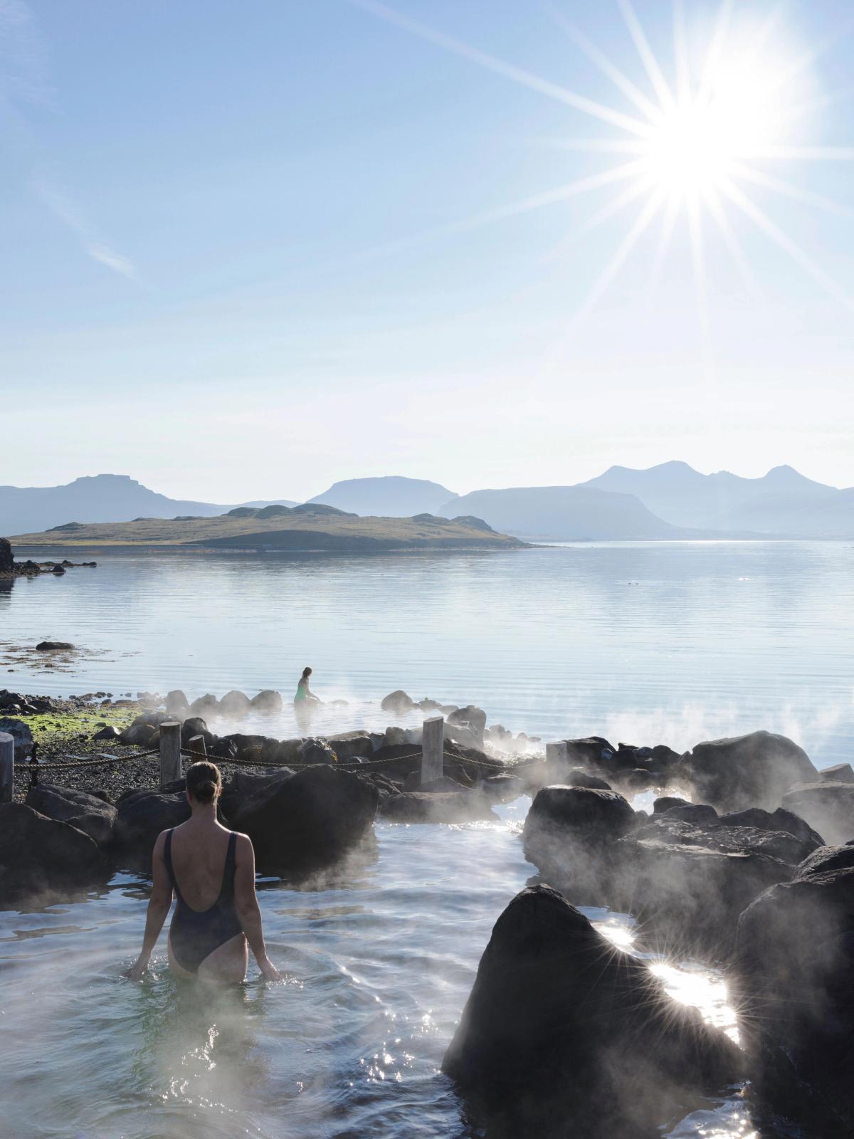 Woman bathing in a steaming geothermal pool next to a lake with mountains under a sunny sky.