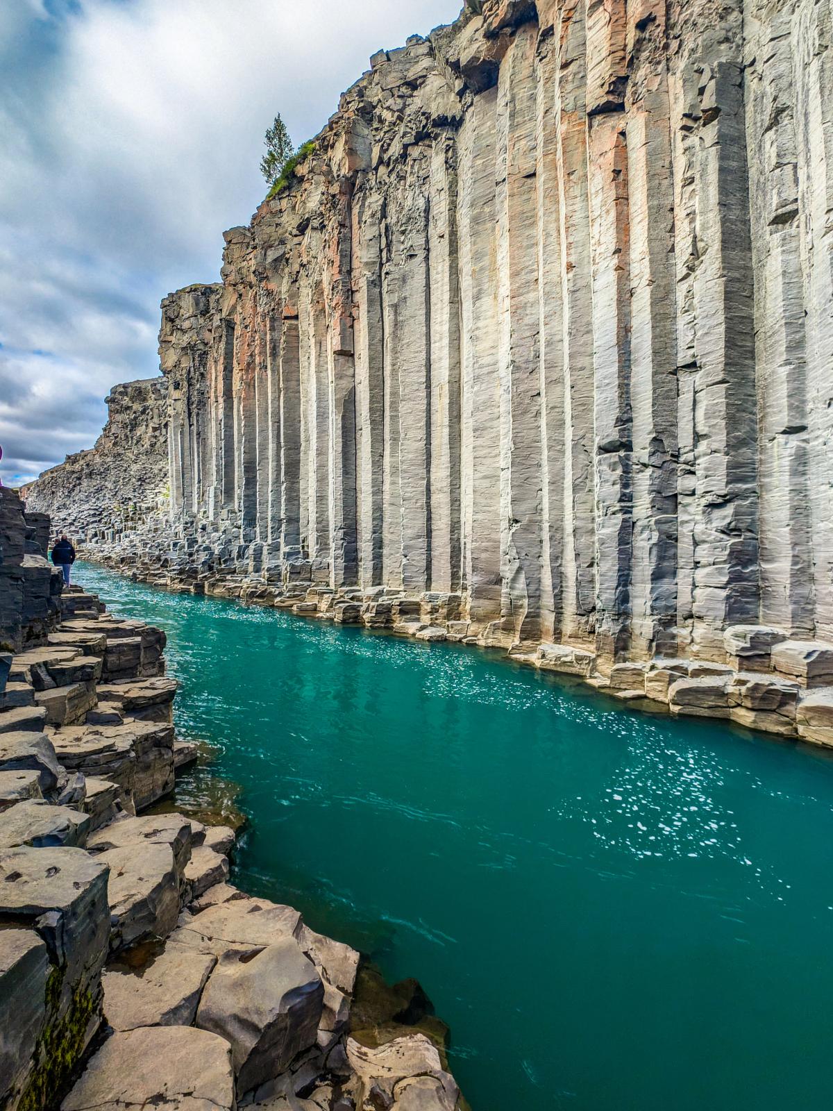 Un río de color turquesa vibrante fluye a través de un cañón con imponentes paredes de basalto columnar, una persona camina por un sendero de formaciones rocosas hexagonales.
