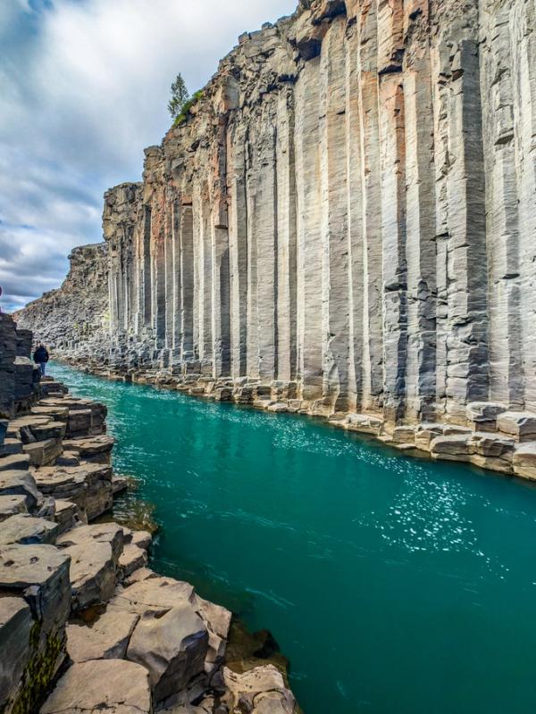 A vibrant turquoise river flows through a canyon with towering columnar basalt walls, a person walks on a path of hexagonal rock formations.