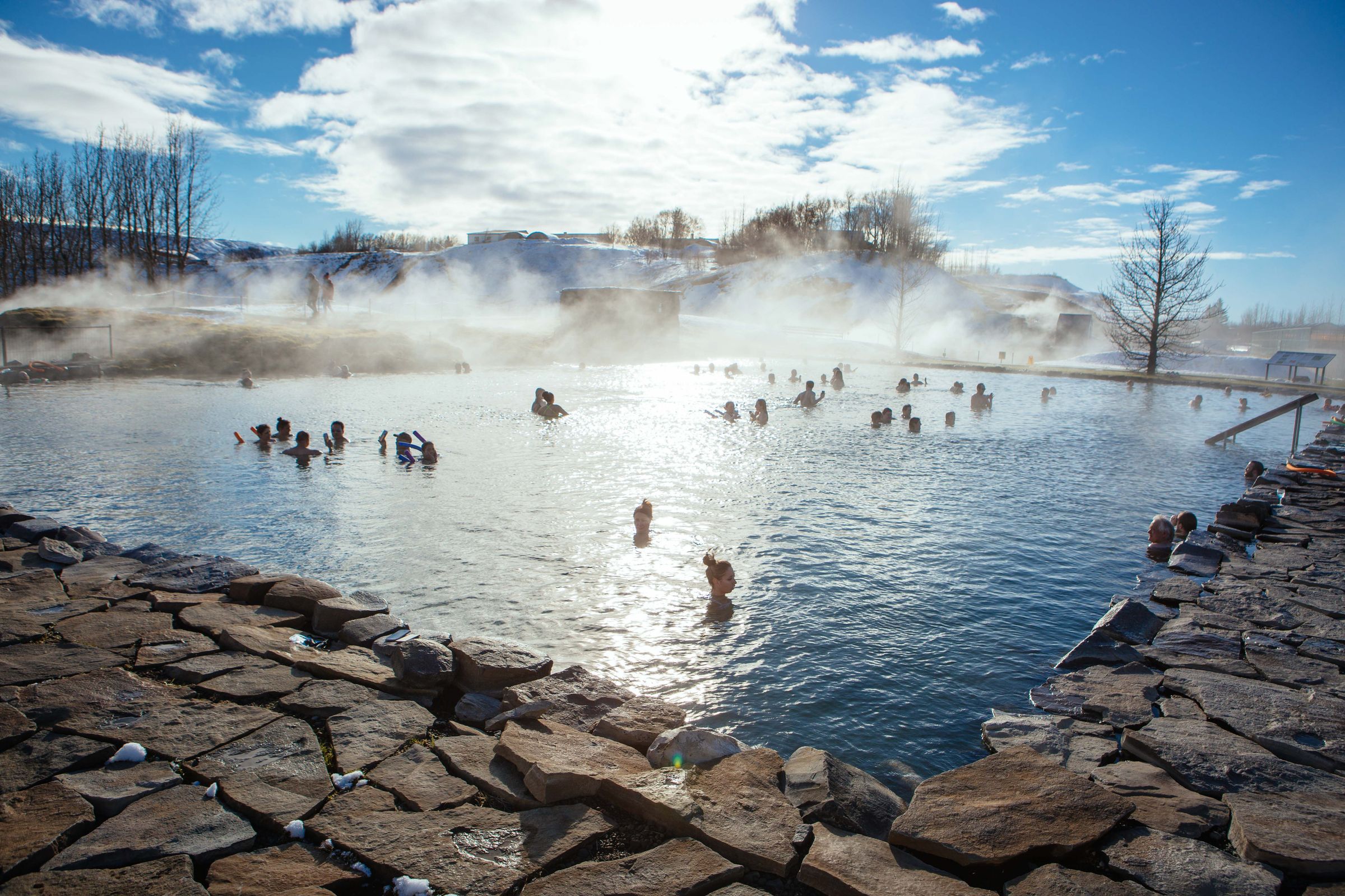 People bathing in a steamy outdoor geothermal hot spring in a snowy landscape.