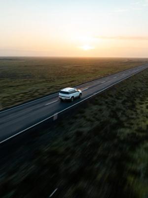 White SUV driving on a long, straight road through a flat, green landscape at sunset.