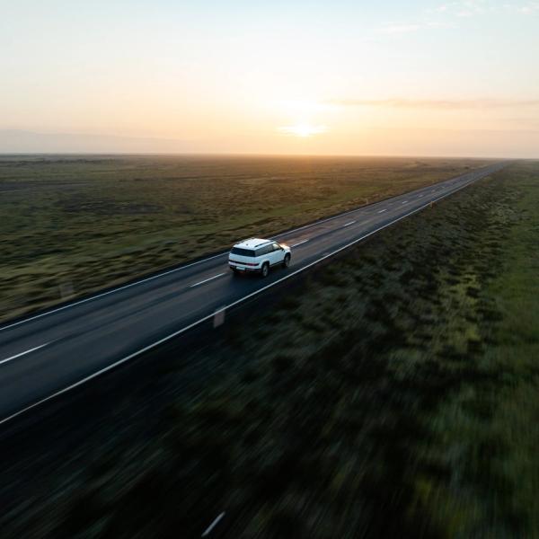 White SUV driving on a long road through a flat, green landscape at sunset.