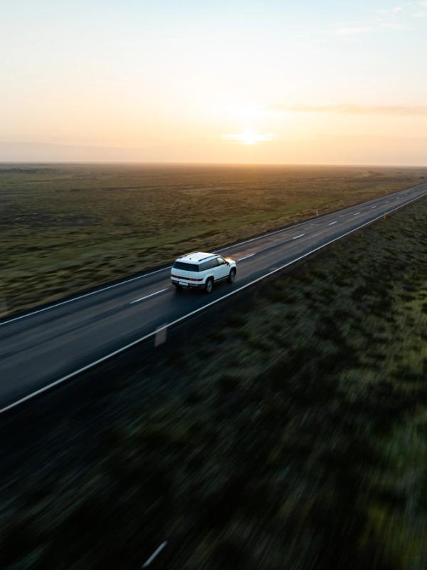 White SUV driving on a long, straight road through a flat, green landscape at sunset.