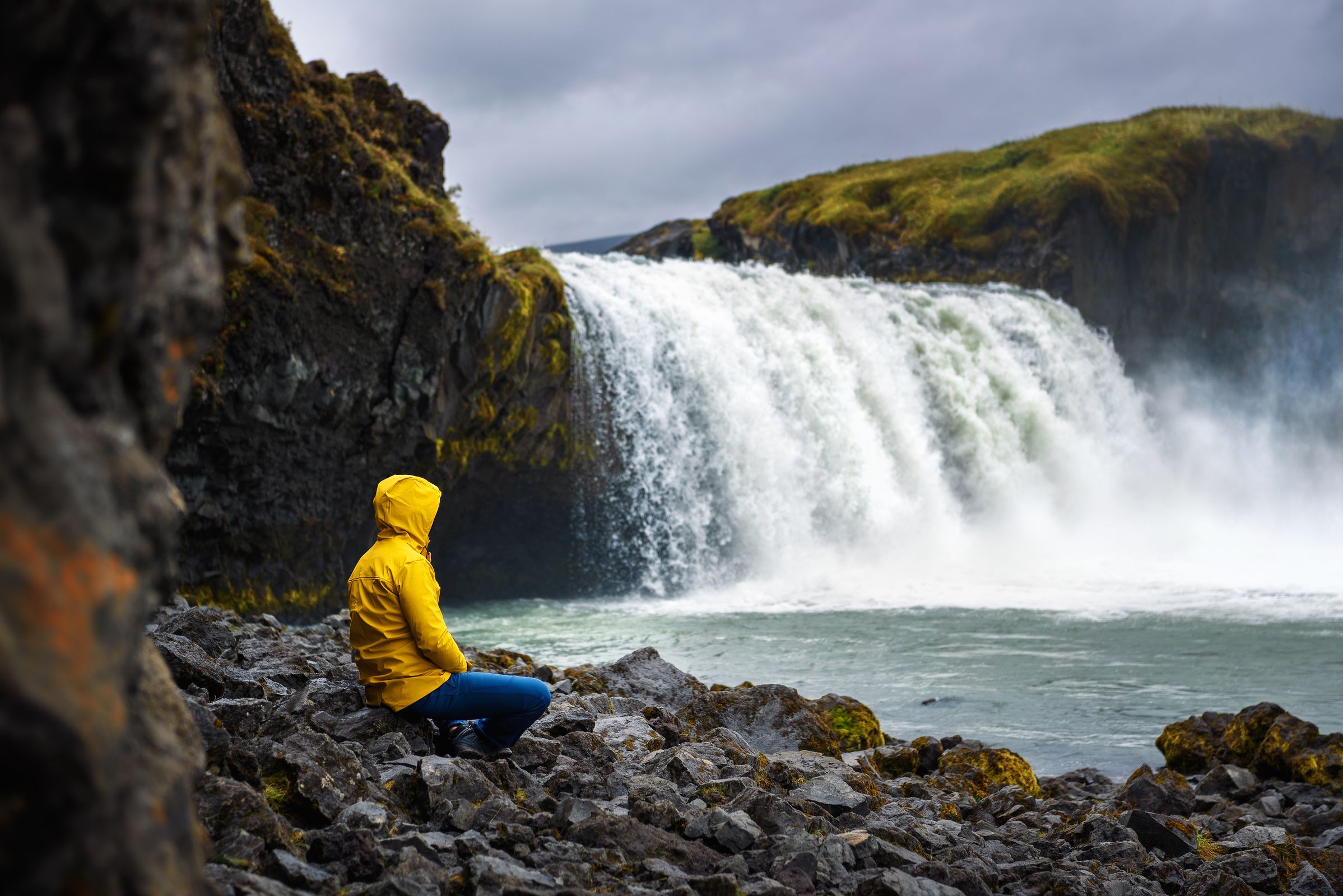 Person in a yellow raincoat squats on a rocky shore facing a large waterfall.