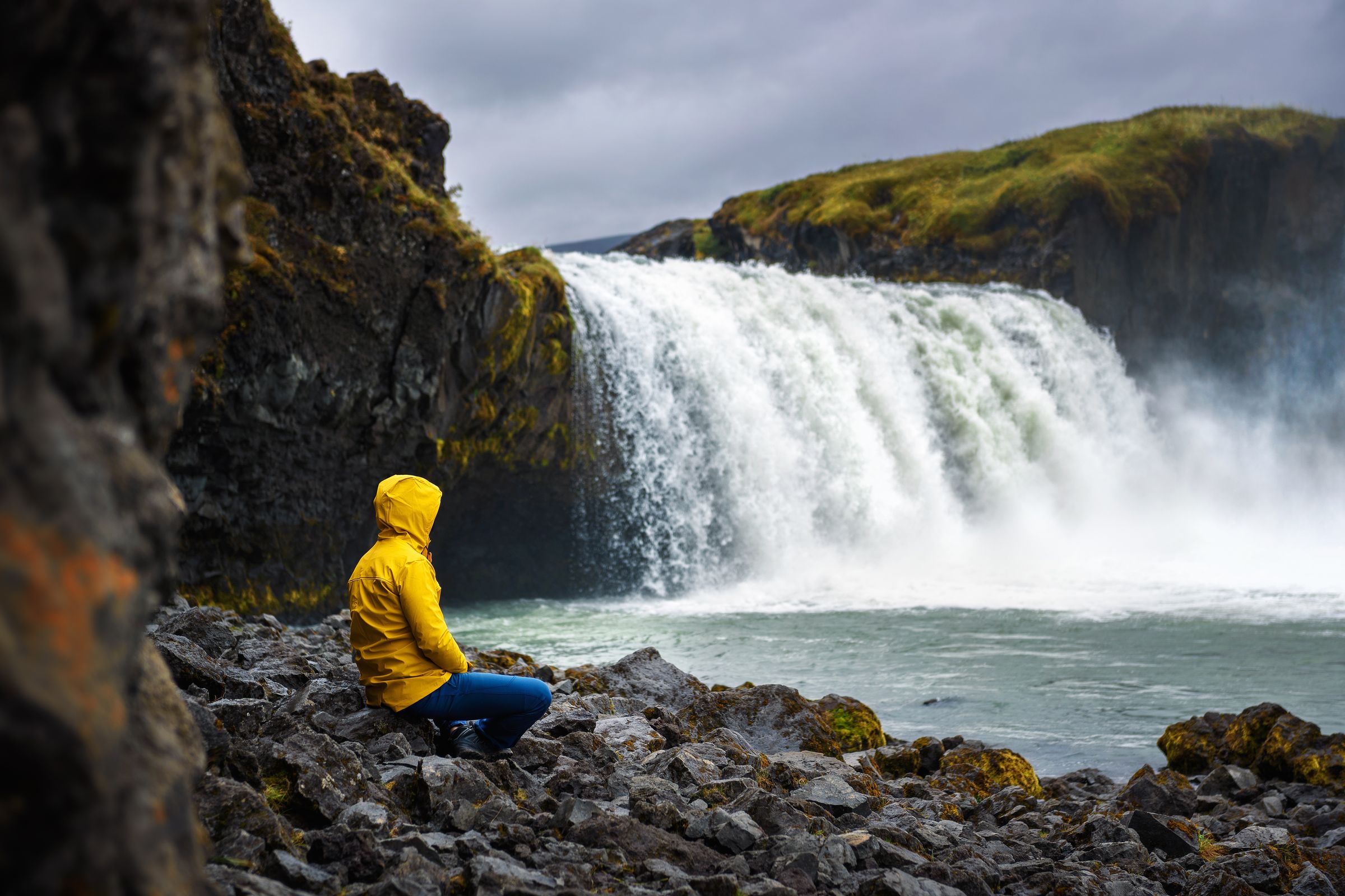 Person in a yellow raincoat squats on a rocky shore facing a large waterfall.
