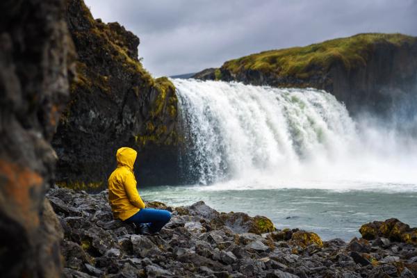a man in a yellow jacket is sitting on the rocks near a waterfall .