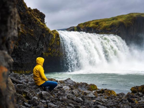 a man in a yellow jacket is sitting on the rocks near a waterfall .