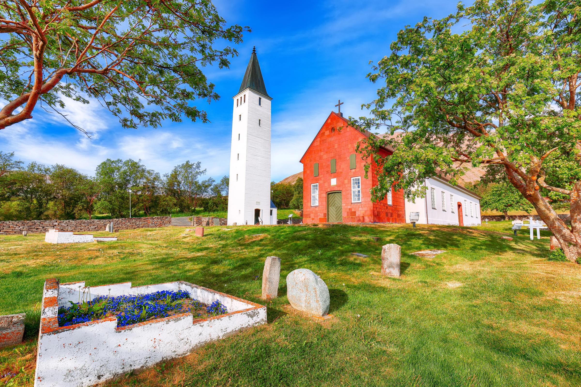 Una iglesia de un rojo intenso con un alto campanario blanco, situada en un cementerio verde bajo un cielo azul brillante.
