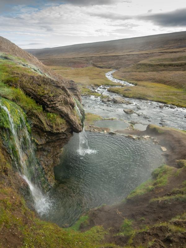 A waterfall cascades over mossy rocks into a natural hot spring pool next to a river in a vast, grassy landscape.